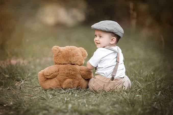 A small child in a flat cap sitting on grass beside a teddy bear.