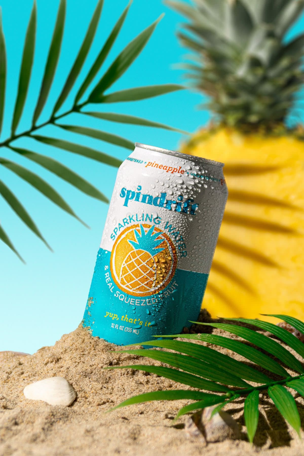 A chilled sparkling-water can styled on sand with palm leaves and a pineapple.
