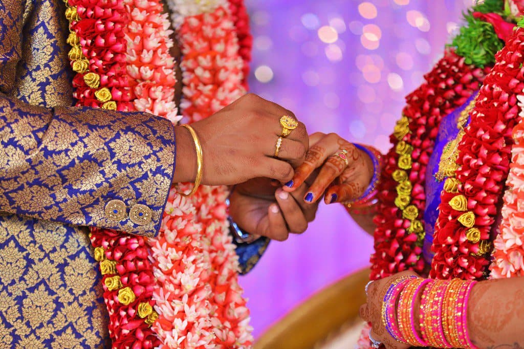 Close detail of a couple's hands during a ring ceremony, with floral garlands behind.