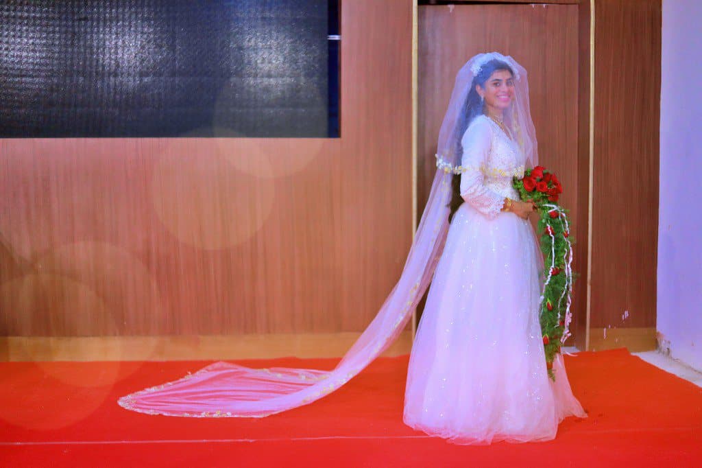 A bride walking a red carpet aisle in a white gown with a long veil.