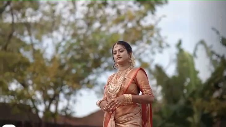 A bride in a gold silk saree and traditional jewellery, filmed outdoors.